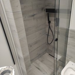 Stol-Lux - Wykończona kabina prysznicowa z czarną armaturą i szarymi płytkami imitującymi drewno, widok z boku na toaletę i fragment narzędzia malarskiego.