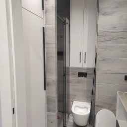 Stol-Lux - Nowoczesna łazienka z szaro-białymi płytkami imitującymi drewno, podwieszana toaleta, białe szafki z czarnymi uchwytami i kabina prysznicowa z czarnymi profilami.