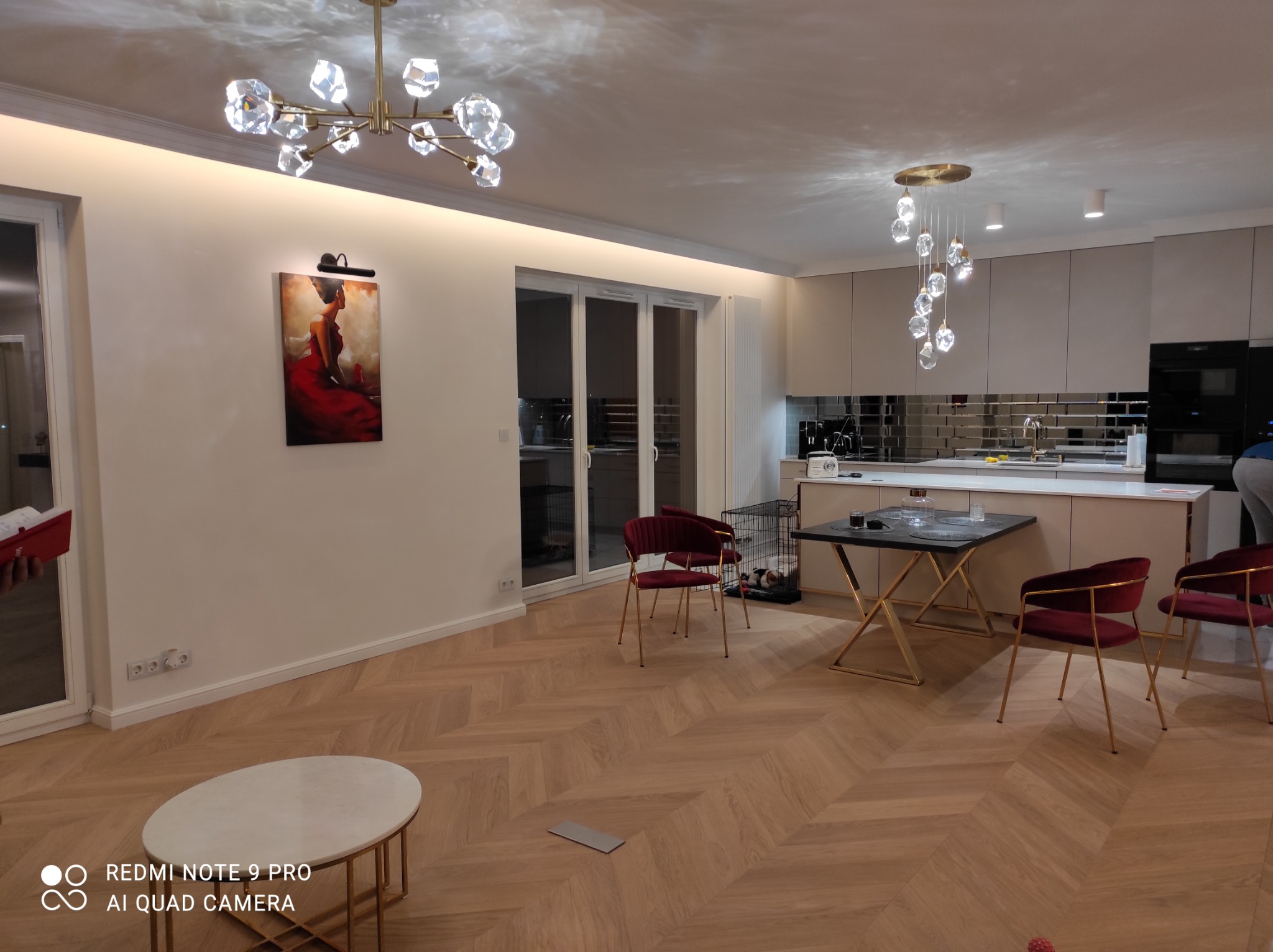 Jasne wnętrze mieszkania z parkietem ułożonym w jodełkę, ozdobnymi lampami sufitowymi, obrazem na ścianie i częściowo widoczną zabudową kuchenną.