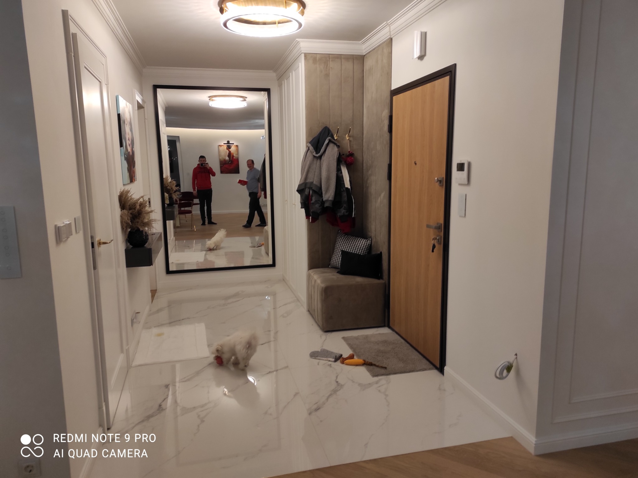 Elegancki przedpokój z marmurową posadzką, dużym lustrem w czarnej ramie odbijającym wnętrze mieszkania, zabudowaną szafą z panelami tapicerowanymi i małym psem bawiącym się na podłodze.