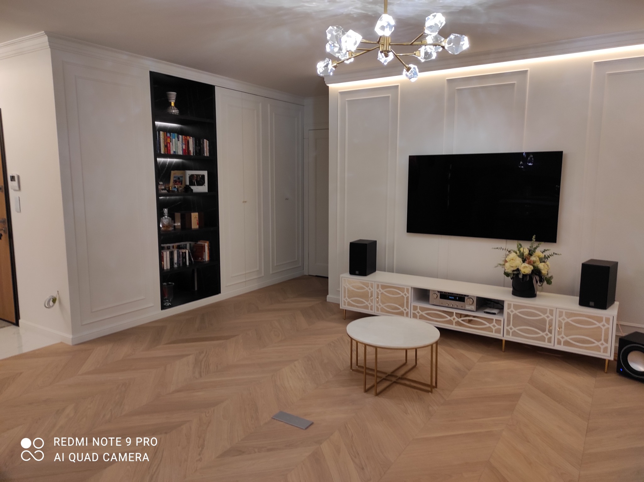 Elegancki salon z parkietem ułożonym w jodełkę, białe ściany z dekoracyjnymi panelami, szafa wnękowa z podświetlanymi półkami, telewizor oraz biała szafka RTV z dekoracyjnymi elementami i okrągły...