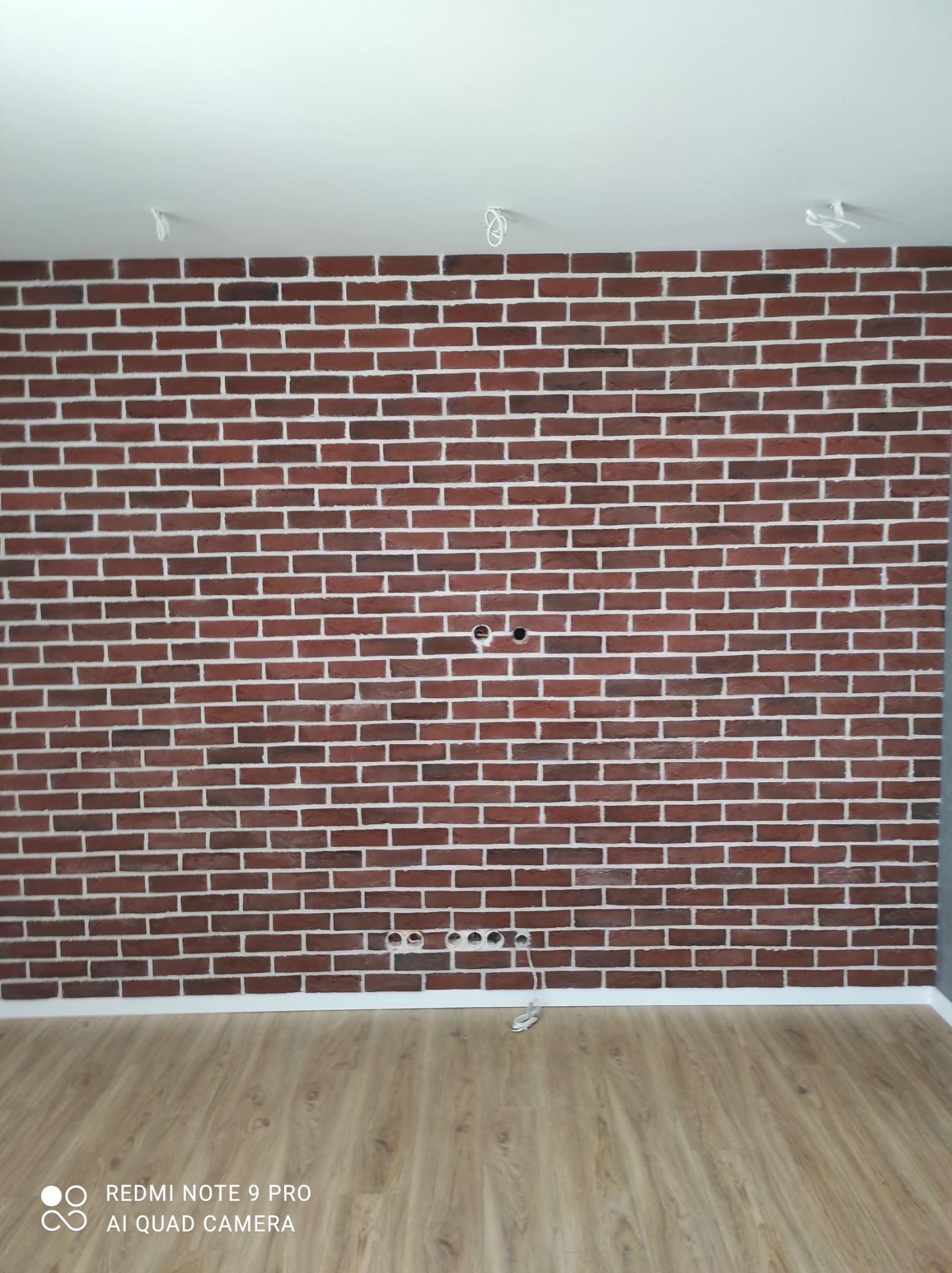 Ściana z cegły klinkierowej w odcieniach czerwieni z białymi fugami, widoczne otwory na gniazdka elektryczne i przewody pod sufitem, panele podłogowe w kolorze jasnego drewna.