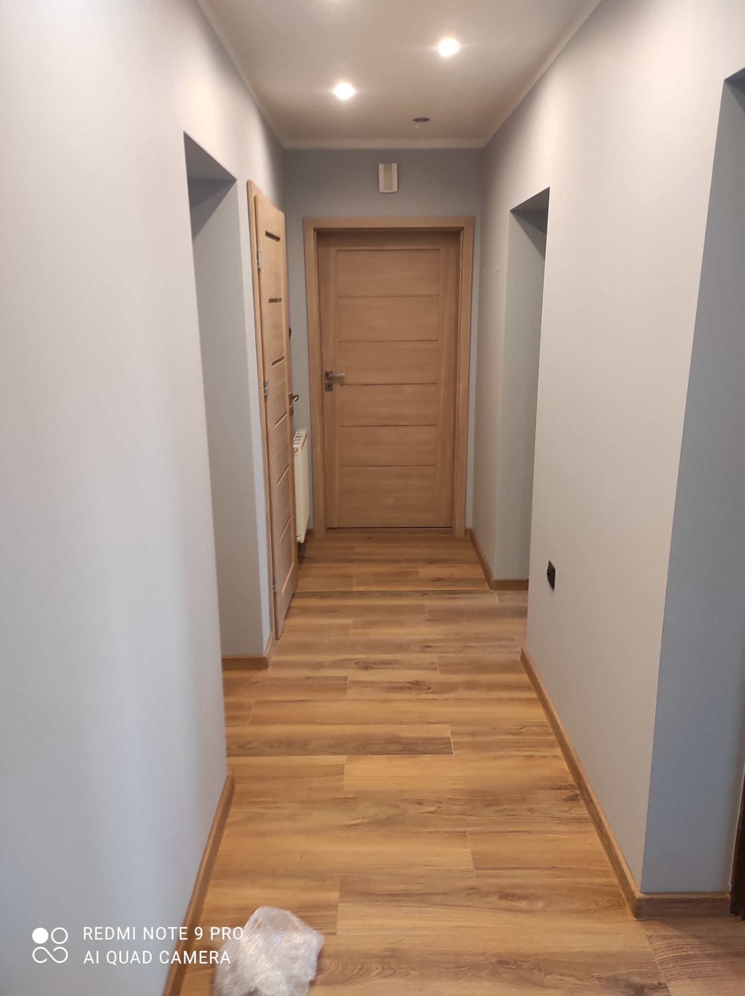 Korytarz po remoncie z panelami podłogowymi w odcieniach brązu, szarymi ścianami i drewnianymi drzwiami. W tle widoczny grzejnik.