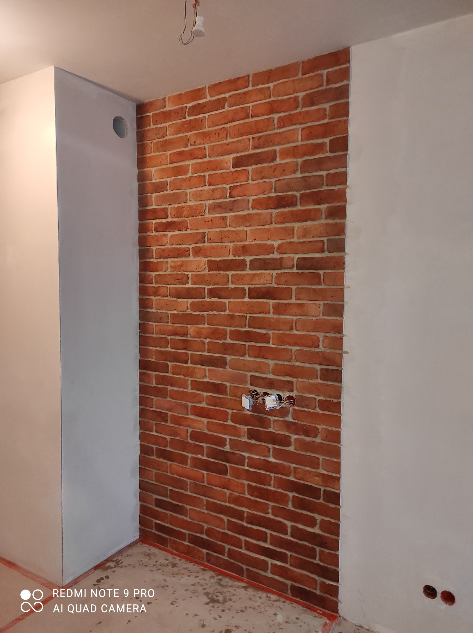 Fragment ściany z dekoracyjną czerwoną cegłą, z widocznymi przewodami elektrycznymi i fragmentem świeżo pomalowanej ściany na biało, w trakcie prac wykończeniowych.