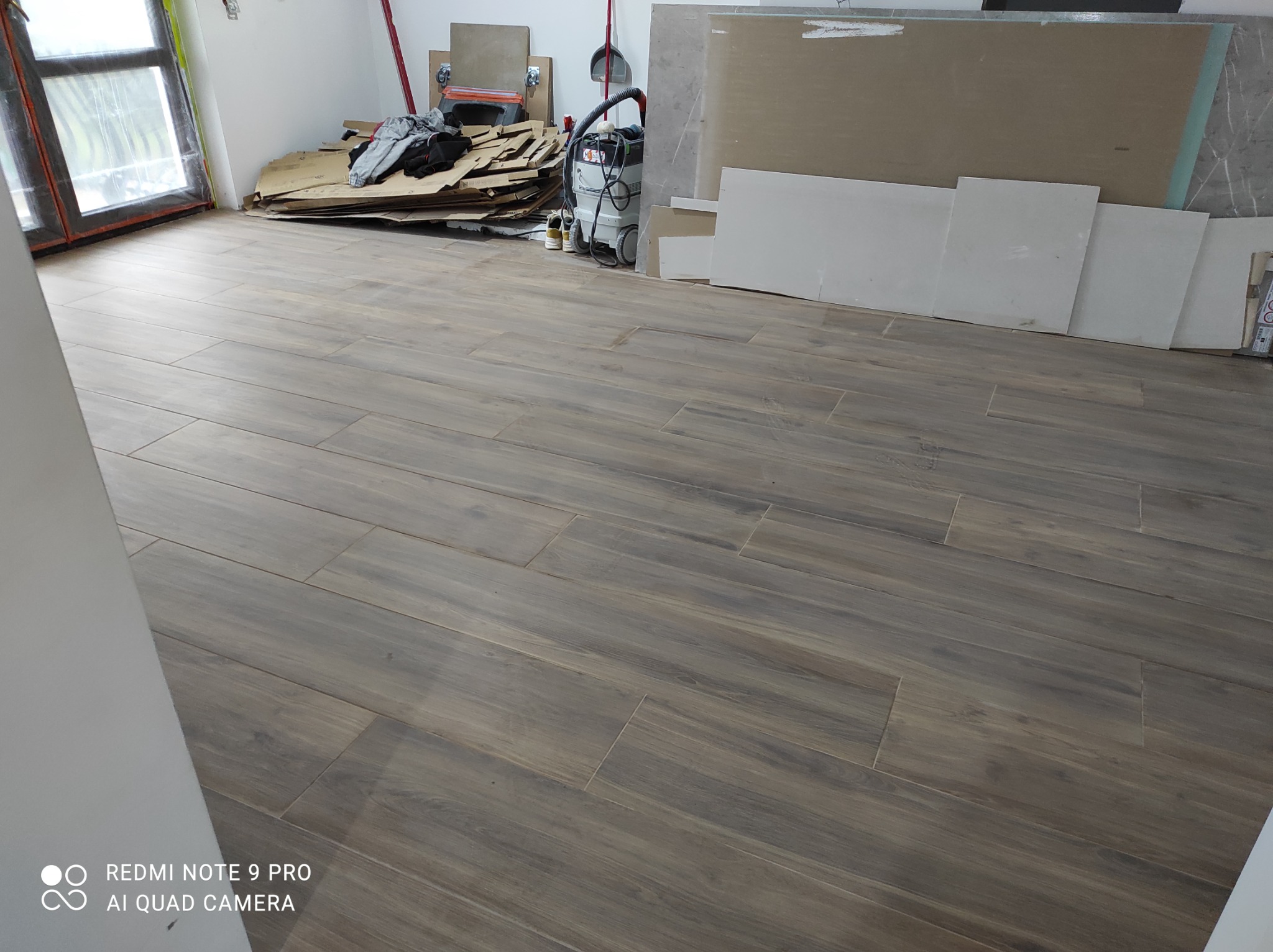 Ułożona podłoga z płytek imitujących drewno w trakcie prac wykończeniowych, widoczne kartony, odkurzacz przemysłowy, płyty gipsowo-kartonowe i resztki materiałów budowlanych.