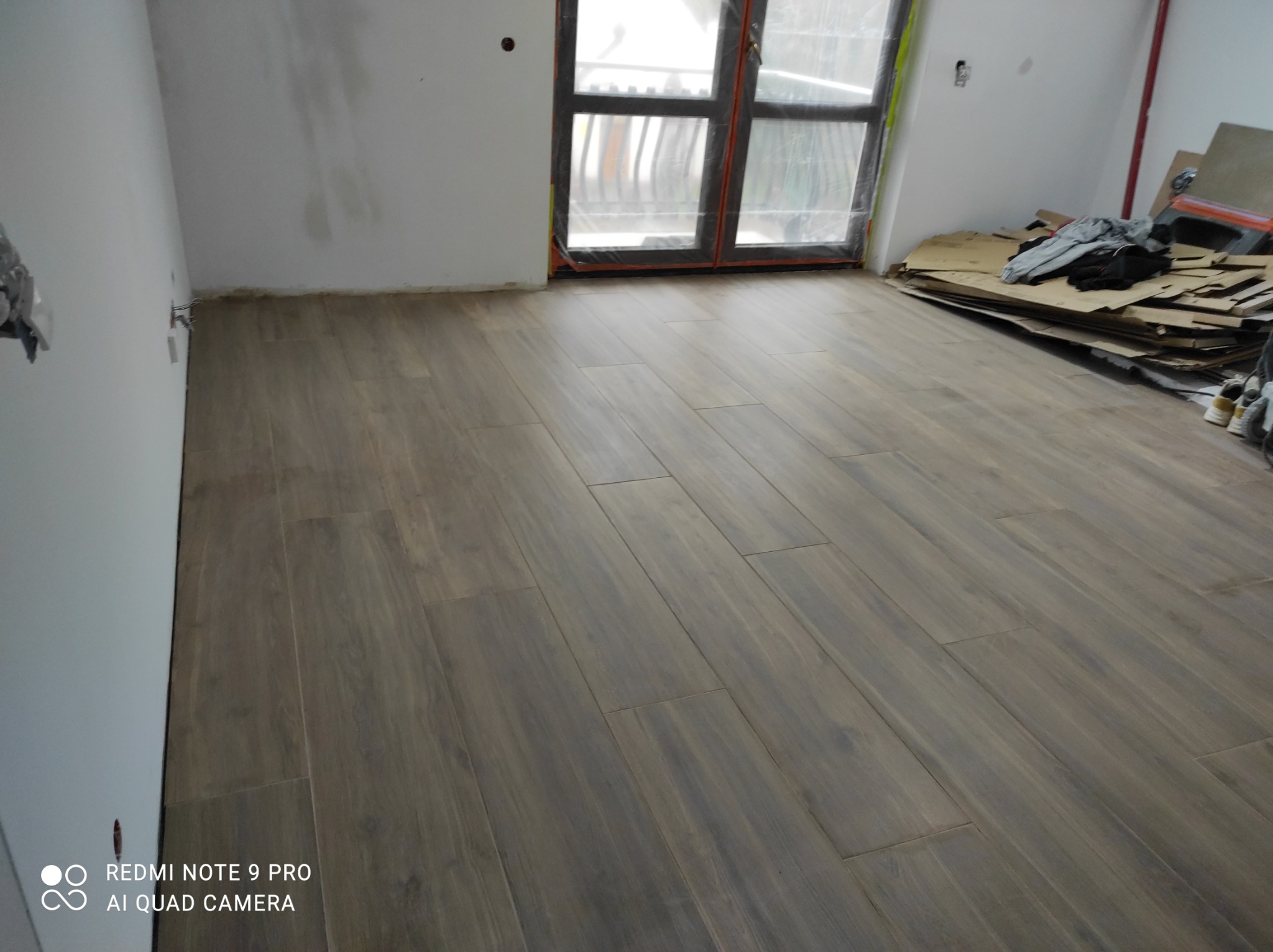 Ułożone panele podłogowe w odcieniach brązu, widoczne przy ścianie niedokończone prace wykończeniowe, okno zabezpieczone folią malarską, kartony i ubrania w rogu pokoju.