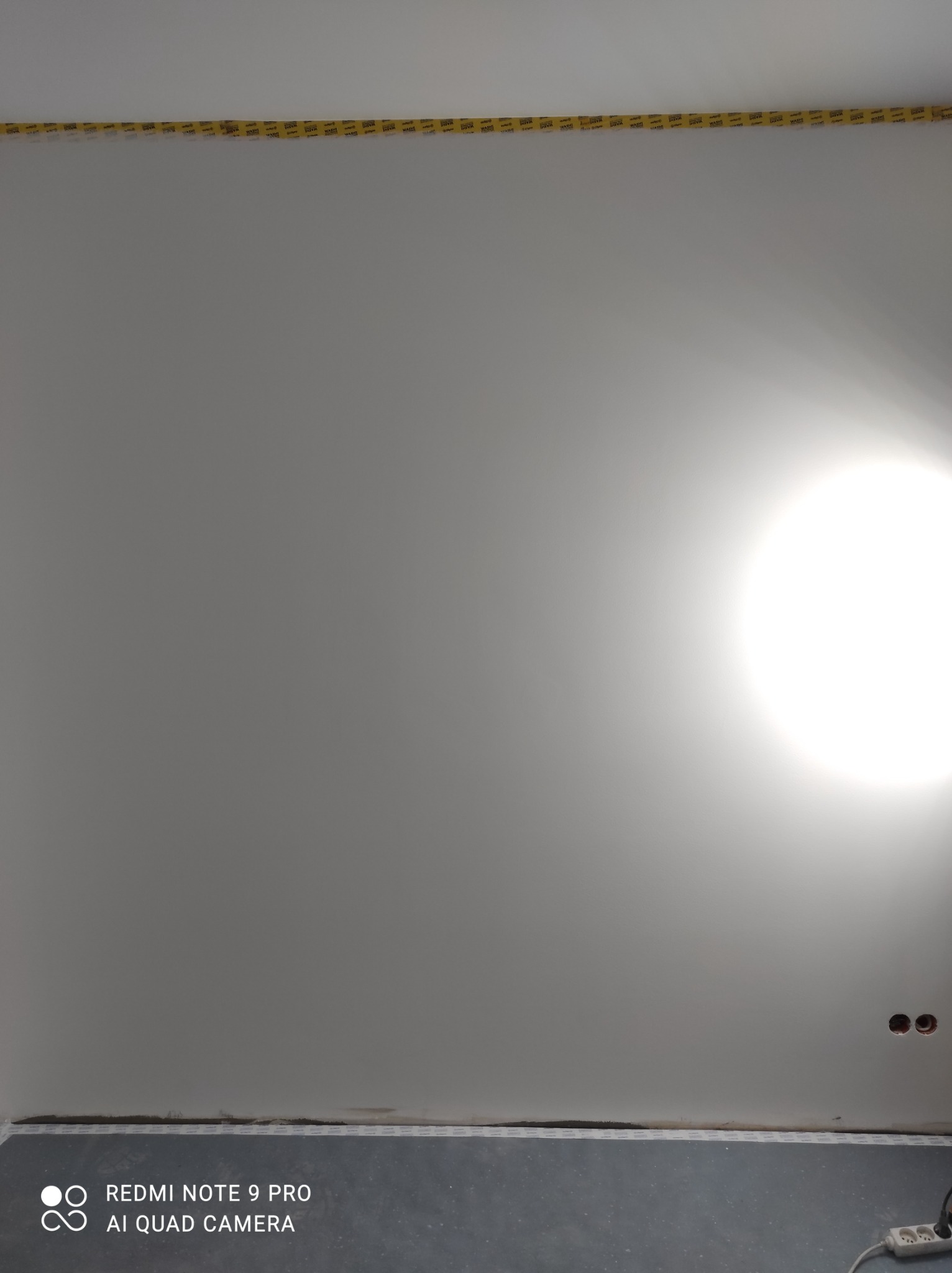 Świeżo wyszpachlowany sufit z taśmą malarską przy krawędzi, widoczne gniazdka elektryczne i fragment podłogi zabezpieczonej folią.