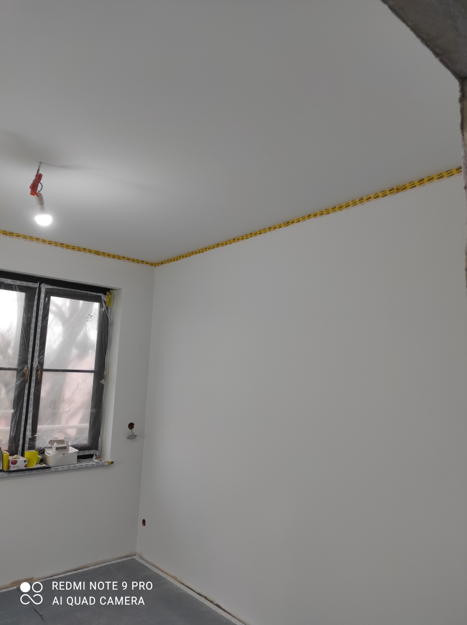 Pokój po szpachlowaniu i malowaniu na biało, zabezpieczony żółtą taśmą malarską przy suficie, okno zakryte folią ochronną.