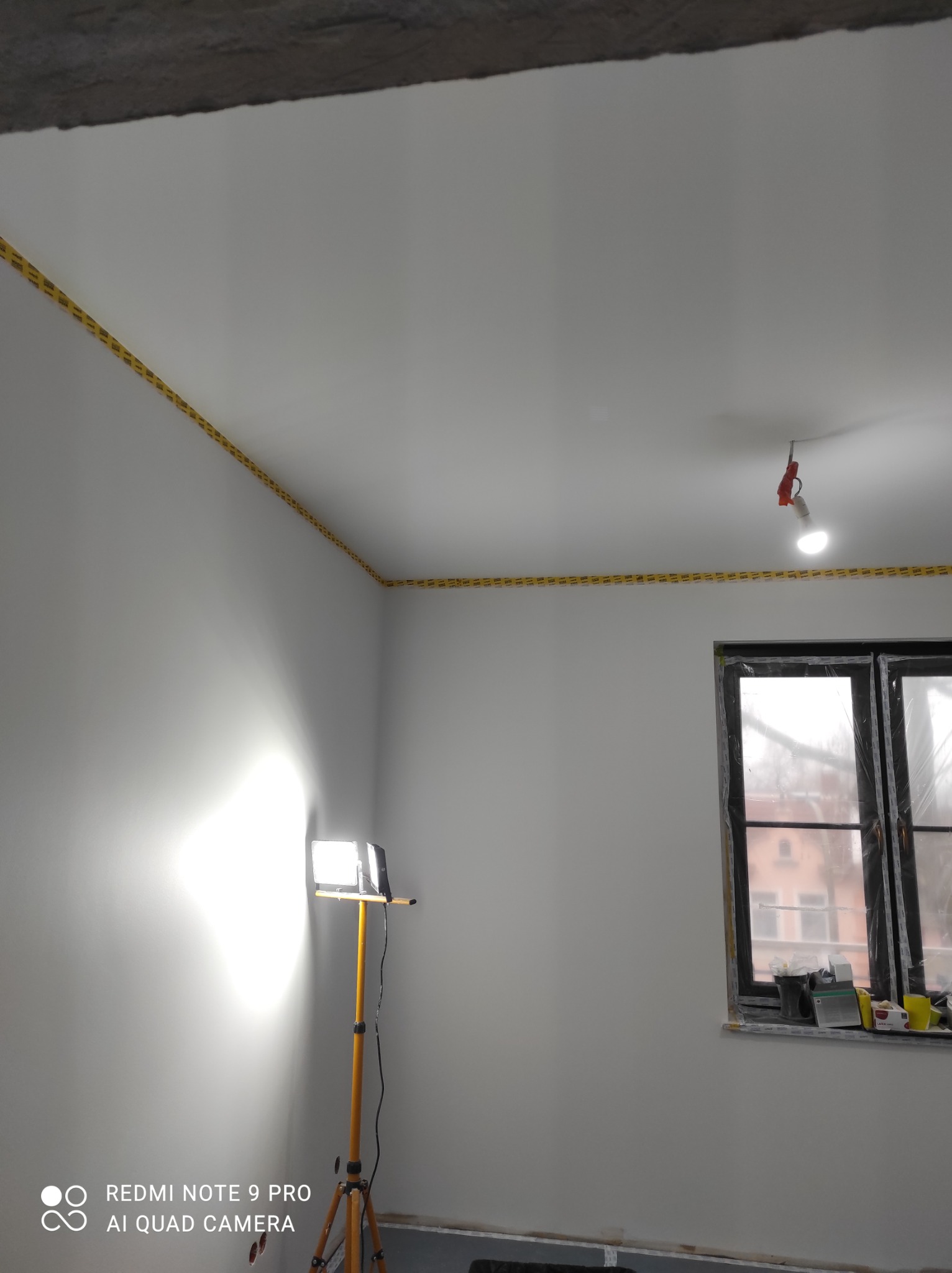 Pokój w trakcie prac wykończeniowych: świeżo pomalowane ściany i sufit z taśmą malarską na krawędziach, okno zabezpieczone folią, halogen na statywie oświetla ścianę.