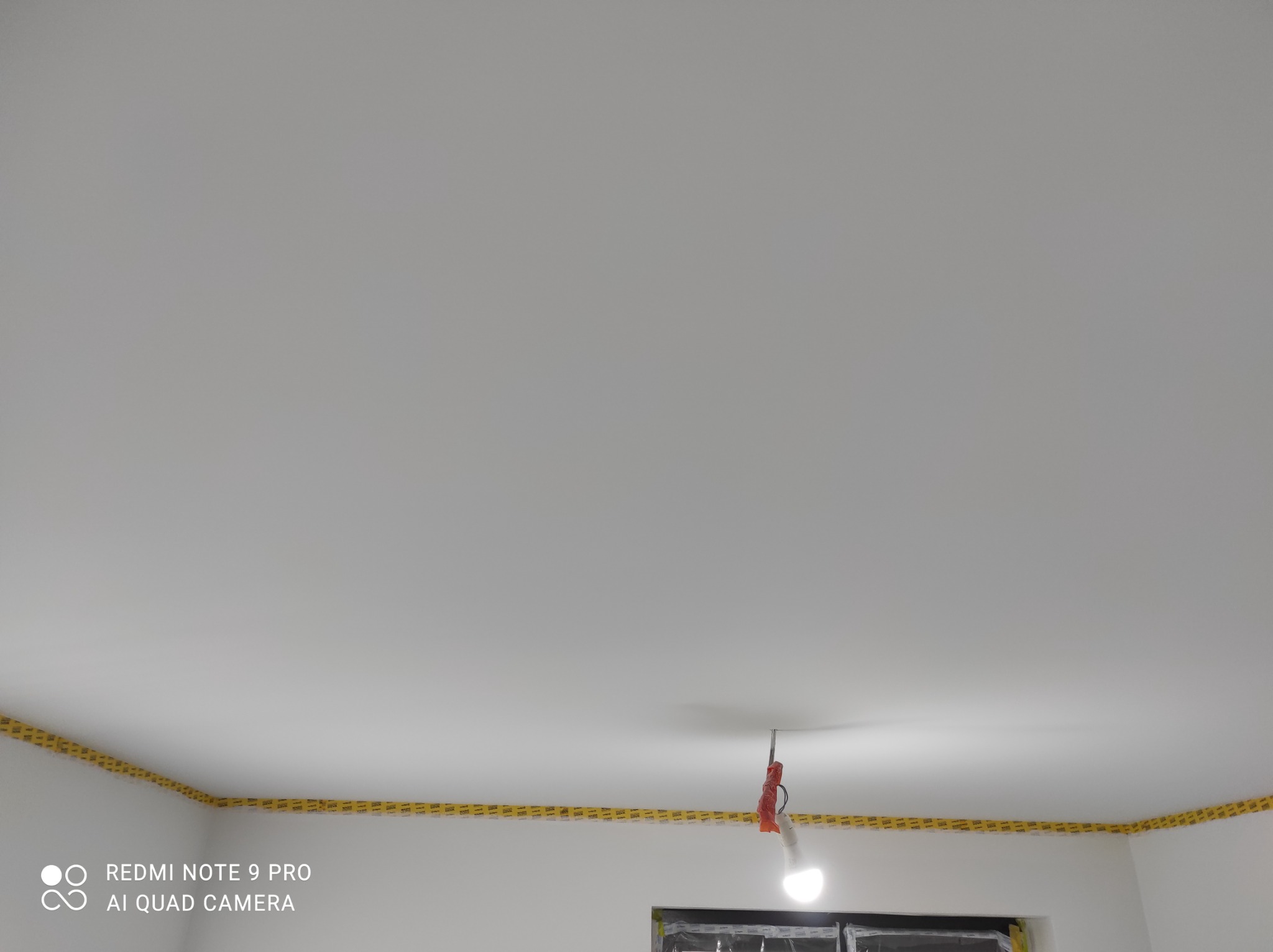 Biały sufit z nałożoną żółtą taśmą malarską wzdłuż krawędzi, lampa tymczasowa zwisa z sufitu, okno zabezpieczone folią.