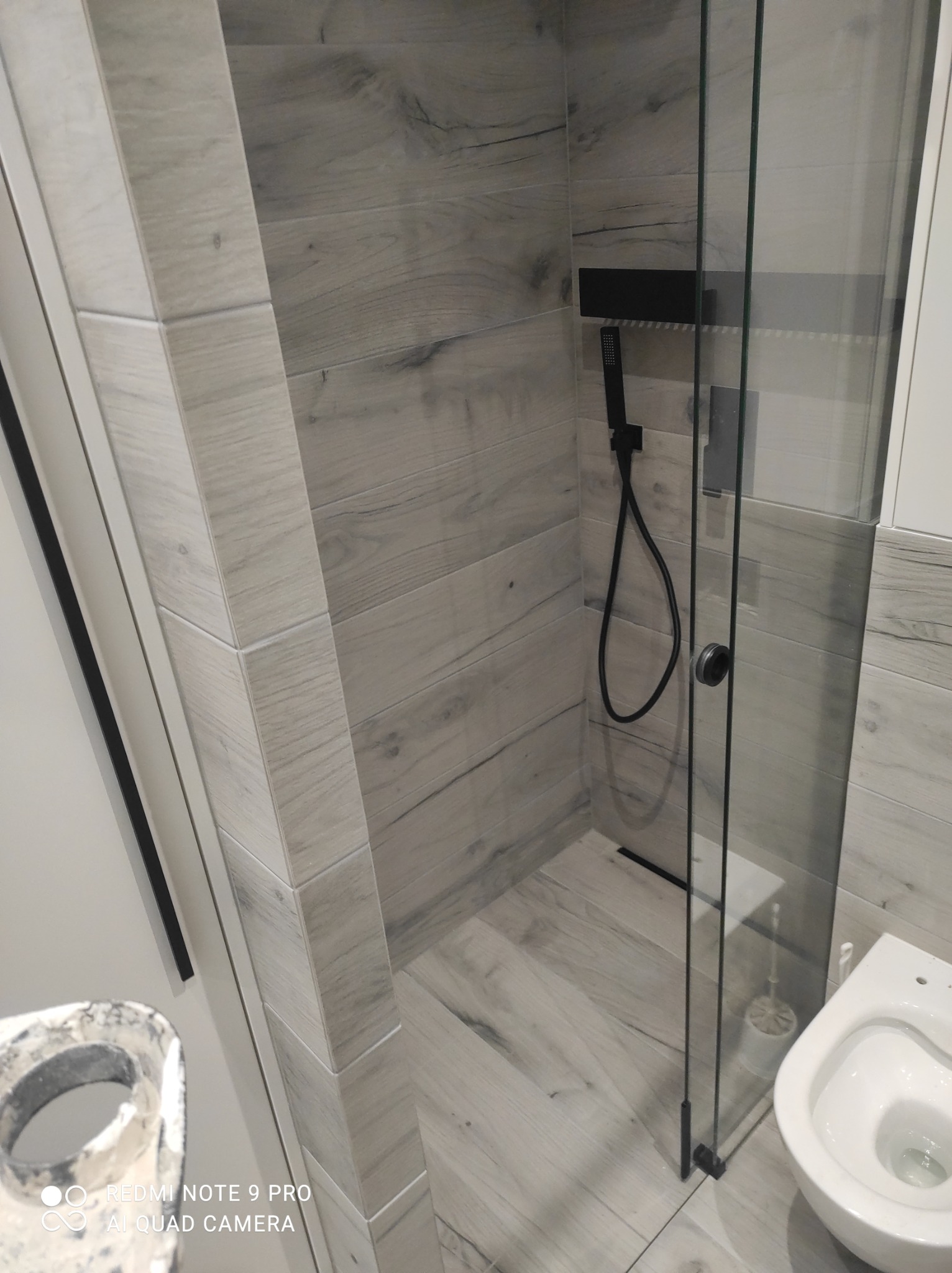 Wykończona kabina prysznicowa z czarną armaturą i szarymi płytkami imitującymi drewno, widok z boku na toaletę i fragment narzędzia malarskiego.