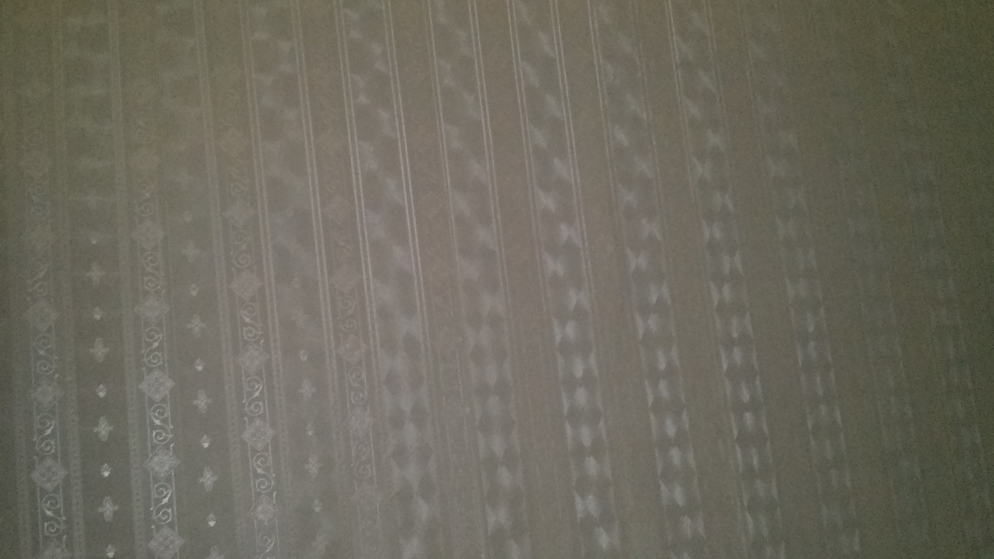 Fragment ściany pokrytej tapetą w odcieniach szarości z delikatnym, pionowym wzorem w paski i motywy roślinne, oświetlonej nierównomiernie.