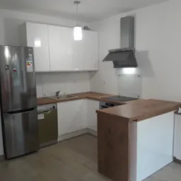 Jasna kuchnia z białymi szafkami, drewnianym blatem i srebrną lodówką LG, widok na wyspę kuchenną i okap.