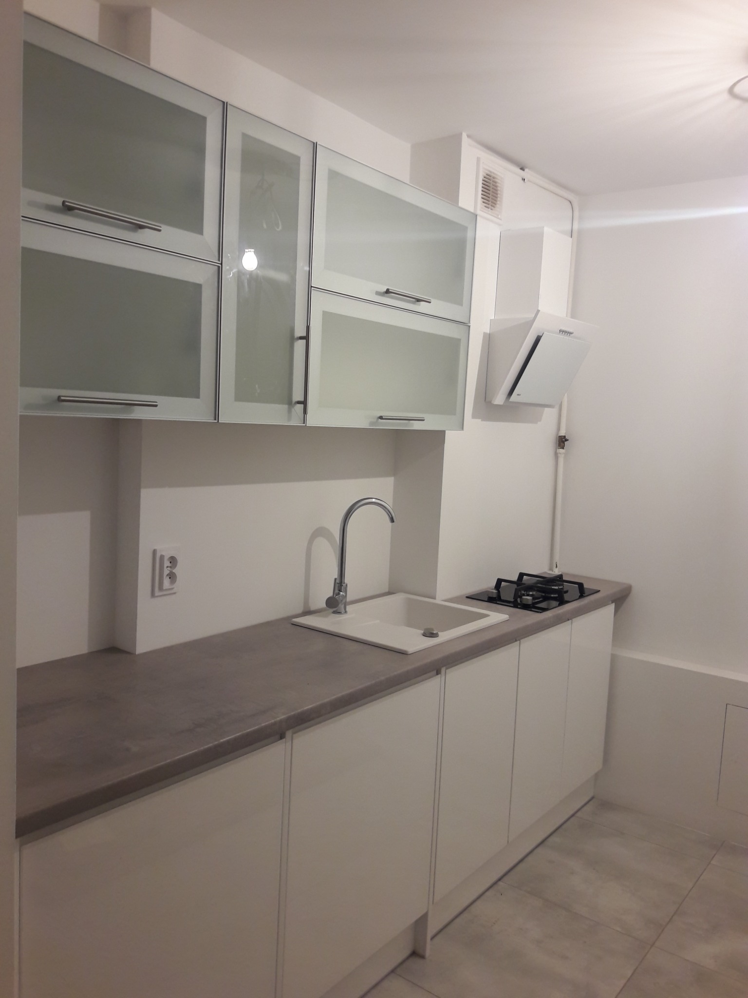 Nowoczesna, minimalistyczna kuchnia z białymi, matowymi frontami szafek, szarym blatem, stalowym zlewem i płytą gazową, oświetlona górnym światłem.