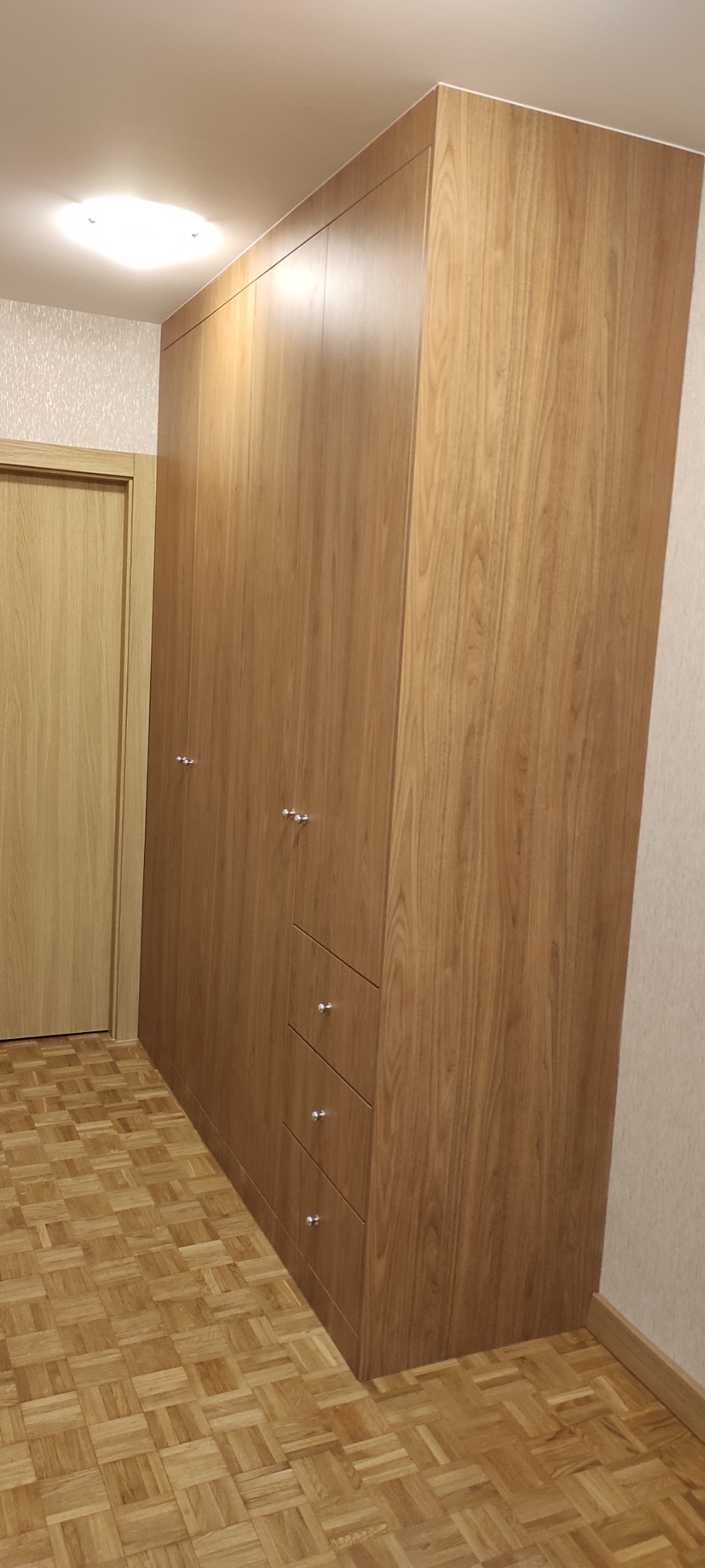 Wysoka, drewniana szafa wnękowa z trzema szufladami i chromowanymi uchwytami, dopasowana do parkietu z jasnego drewna, widoczna obok jasnych, drewnianych drzwi.