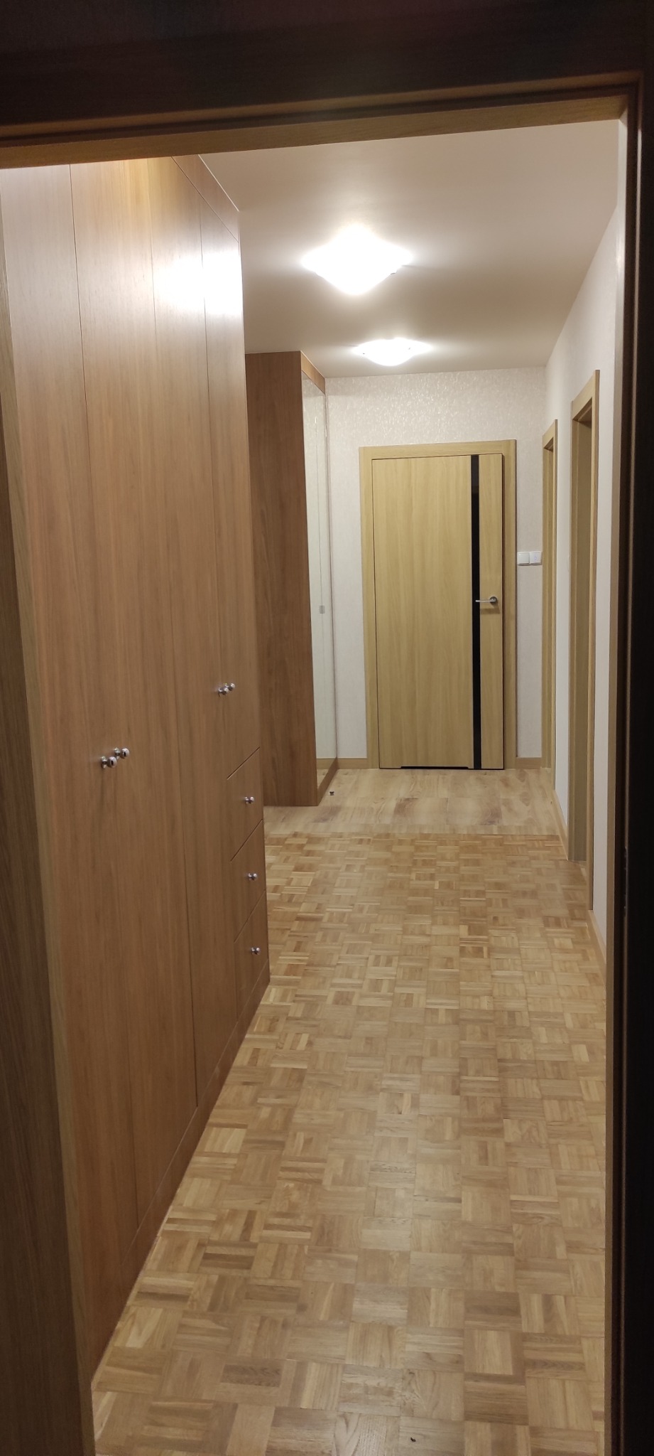 Wnętrze korytarza z zabudowaną szafą wnękową z jasnego drewna, parkietem w kwadraty i drzwiami z czarnym pasem.