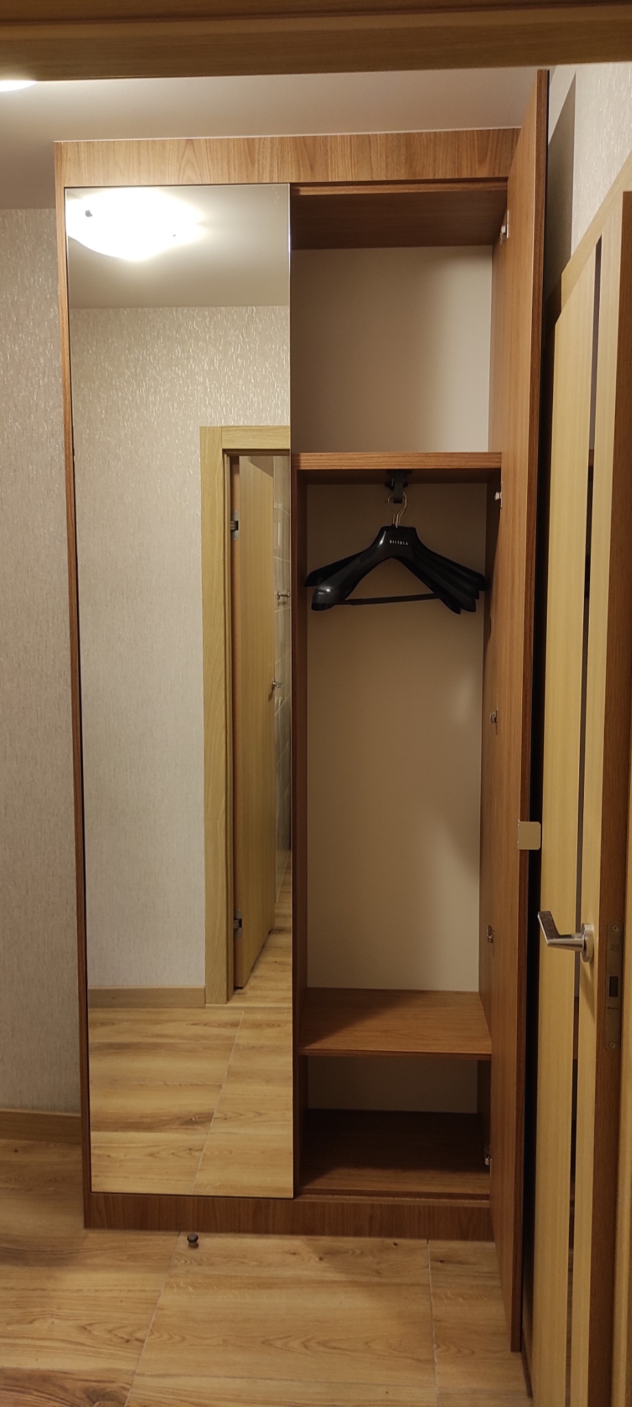 Wysoka szafa z lustrem i otwartą przestrzenią z wieszakiem, widok na korytarz w odbiciu lustra, drewniane wykończenie.