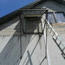 Szklane rury kolektora słonecznego zamontowane na metalowej konstrukcji przy ścianie budynku z szarego bloczka, widoczna drabina oparta o ścianę i cień rzucany przez konstrukcję.