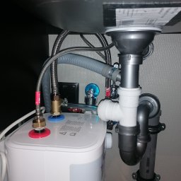 Kamil Brejnak - Instalacja pod zlewem: bojler elektryczny podłączony do rur odpływowych i zaworów, widoczne wężyki doprowadzające wodę oraz elementy mocujące.