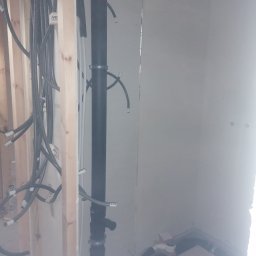 Kamil Brejnak - Surowe wnętrze łazienki w trakcie remontu: widoczne pionowe rury kanalizacyjne z obejmami, szare peszle instalacyjne wyprowadzone ze ściany z drewnianych belek oraz fragment podłogi z odpływem.