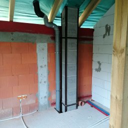 Kamil Brejnak - Wnętrze w trakcie budowy: pion kanalizacyjny z szarej rury, zabezpieczony czarną ramą, przechodzi przez otwór w surowej podłodze; widoczne instalacje elektryczne i fragmenty ścian z różnych...