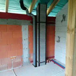 Wnętrze w trakcie budowy: pion kanalizacyjny z szarej rury, zabezpieczony czarną ramą, przechodzi przez otw&oacute;r w surowej podłodze; widoczne instalacje elektryczne i fragmenty ścian z r&oacute;żnych...