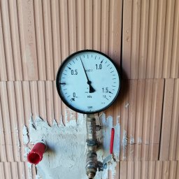 Kamil Brejnak - Manometr zamontowany na instalacji wodnej, widoczny na tle ściany z pustaków ceramicznych, z fragmentami zaprawy wokół rur.