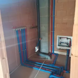 Instalacja wodna i kanalizacyjna w trakcie montażu w surowym pomieszczeniu, widoczne rury w kolorze niebieskim i czerwonym, mocowane do ściany z pustaków ceramicznych, szara rura kanalizacyjna...