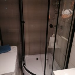 Kamil Brejnak - Narożna kabina prysznicowa z czarnymi profilami w małej łazience, obok pralka i umywalka, widoczny fragment instalacji wodnej.