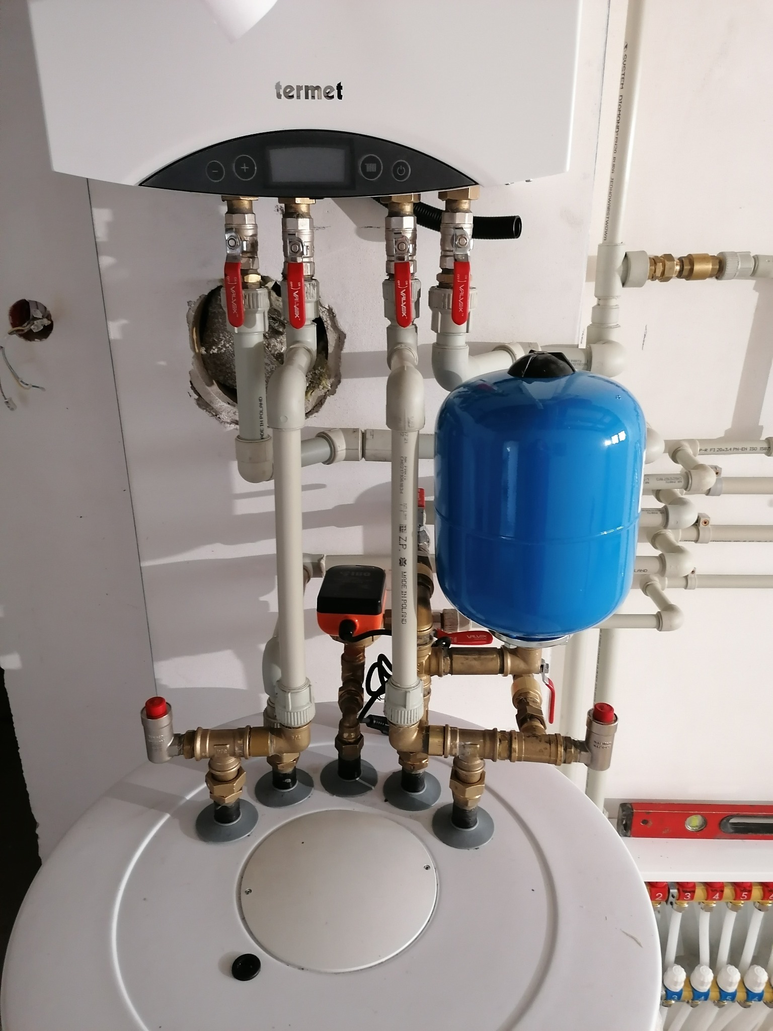 Zamontowany biały piec gazowy Termet z podłączeniami hydraulicznymi, niebieski zbiornik wyrównawczy i rozdzielacz ogrzewania podłogowego z zaworami, widoczny poziomica w tle.
