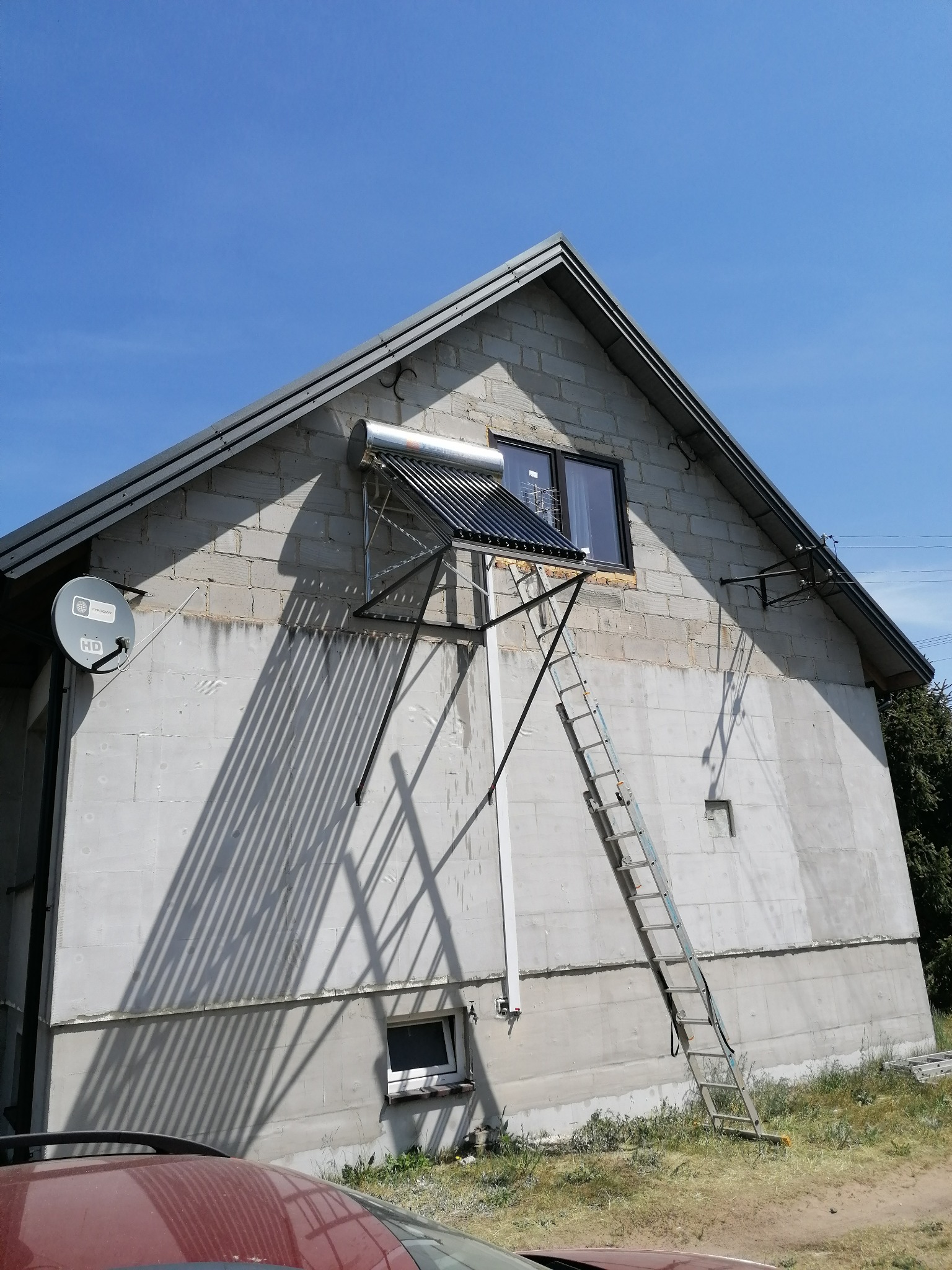 Zewnętrzny montaż kolektora słonecznego na ścianie domu z bloków, widoczna drabina oparta o ścianę, cień rzucany przez kolektor, antena satelitarna z boku.