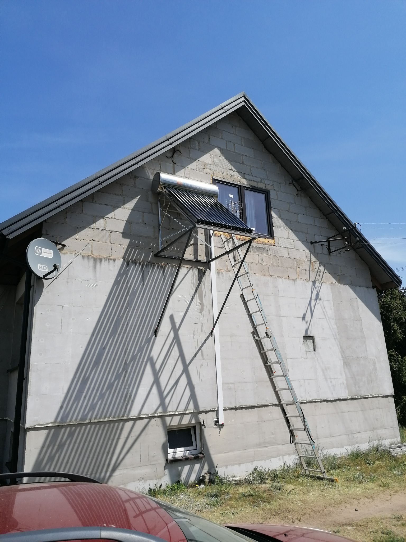 Zewnętrzny kolektor słoneczny zamontowany na ścianie budynku z bloczków betonowych, drabina oparta o ścianę, antena satelitarna na elewacji, widoczne okno i fragment dachu.