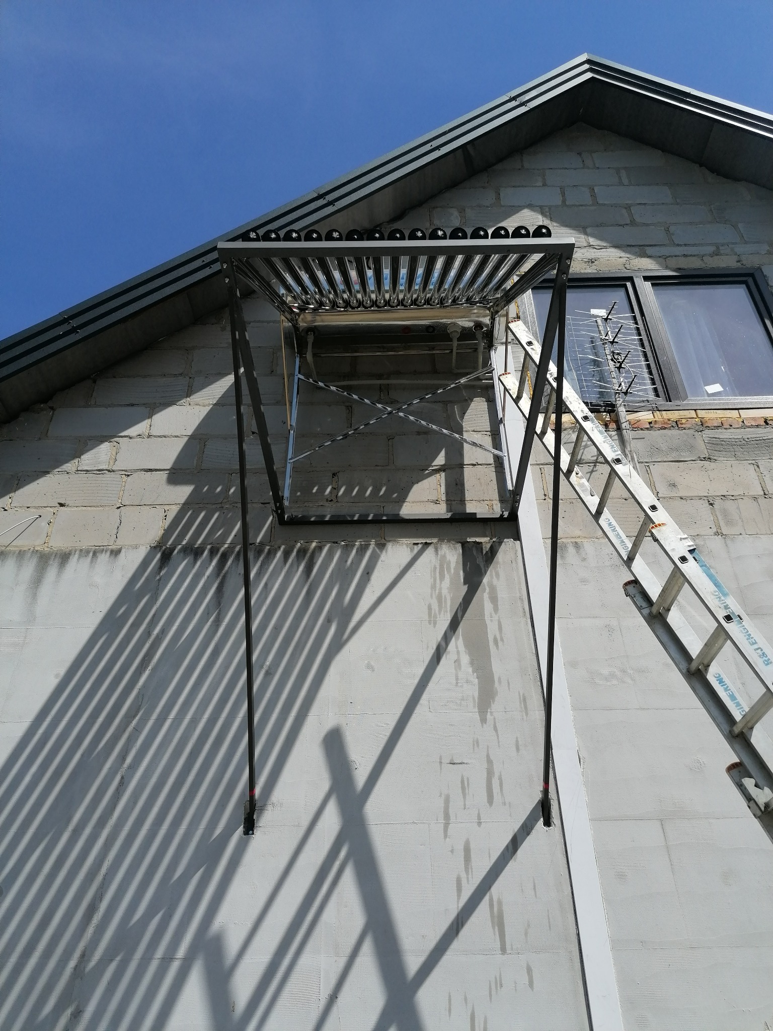 Szklane rury kolektora słonecznego zamontowane na metalowej konstrukcji przy ścianie budynku z szarego bloczka, widoczna drabina oparta o ścianę i cień rzucany przez konstrukcję.