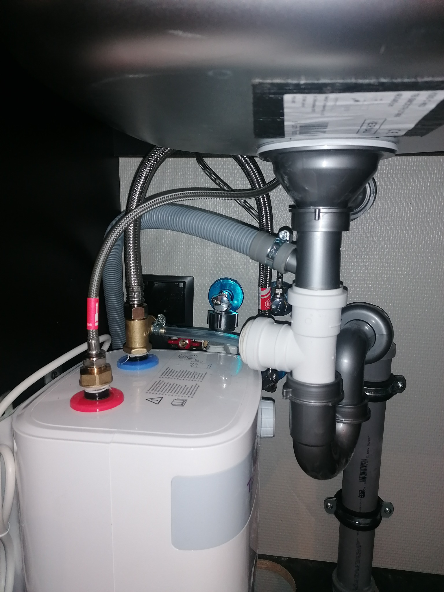 Instalacja pod zlewem: bojler elektryczny podłączony do rur odpływowych i zaworów, widoczne wężyki doprowadzające wodę oraz elementy mocujące.