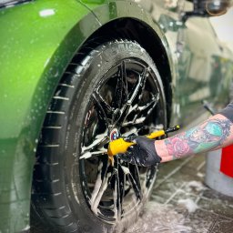Hills detailing and SPA - Auto detailing Poznań, Powłoka ceramiczna, Folia PPF - Zielony samochód podczas mycia felg, widoczna ręka w czarnej rękawicy z tatuażem, używająca szczotki do czyszczenia felg.