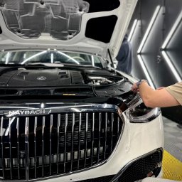 Hills detailing and SPA - Auto detailing Poznań, Powłoka ceramiczna, Folia PPF - Biały Maybach z otwartą maską, w trakcie zabezpieczania folią ochronną przedniej lampy przez pracownika w warsztacie detailingowym z oświetleniem LED.