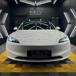 Hills detailing and SPA - Auto detailing Poznań, Powłoka ceramiczna, Folia PPF - Biała Tesla Model 3 po zabiegach pielęgnacyjnych w sterylnym, oświetlonym studiu detailingowym z logo firmy na tablicy rejestracyjnej i podłodze w żółto-czarną kratę.