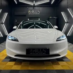 Biała Tesla Model 3 po zabiegach pielęgnacyjnych w sterylnym, oświetlonym studiu detailingowym z logo firmy na tablicy rejestracyjnej i podłodze w żółto-czarną kratę.