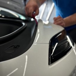 Hills detailing and SPA - Auto detailing Poznań, Powłoka ceramiczna, Folia PPF - Aplikacja folii ochronnej na przednią część białego samochodu, widoczny fragment reflektora i narzędzie do aplikacji w ręce pracownika.