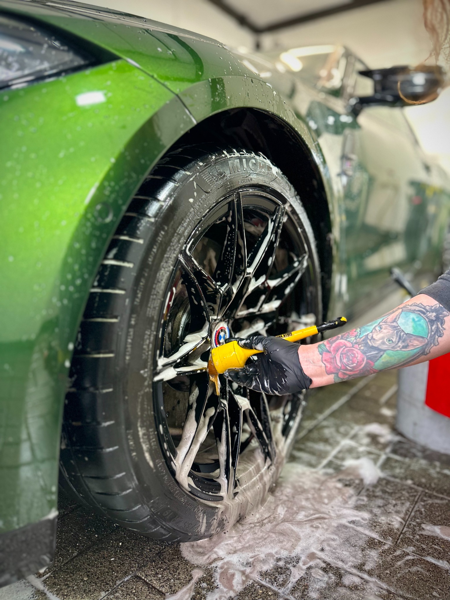 Zielony samochód podczas mycia felg, widoczna ręka w czarnej rękawicy z tatuażem, używająca szczotki do czyszczenia felg.
