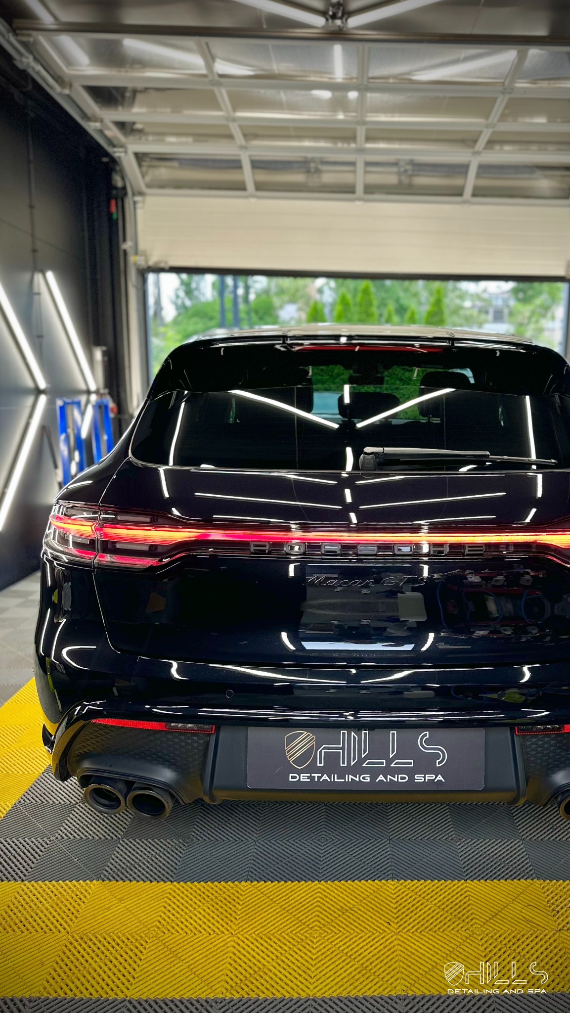 Błyszczący, czarny Porsche Macan GTS po zabiegach pielęgnacyjnych, sfotografowany od tyłu w dobrze oświetlonym garażu z widocznym logo firmy detailingowej.