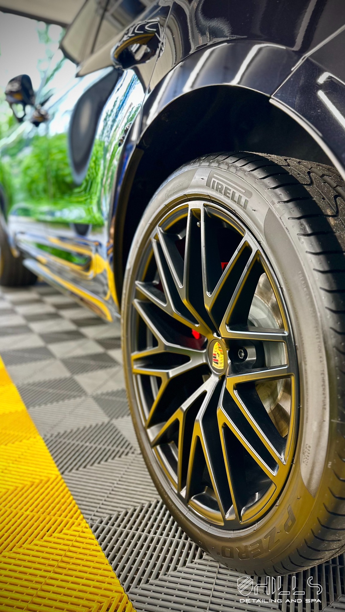 Błyszczące, czarne Porsche Macan na podłodze w kratkę podczas detailingu, widoczne odbicie w lakierze, felga z logo Porsche i opona Pirelli P Zero.