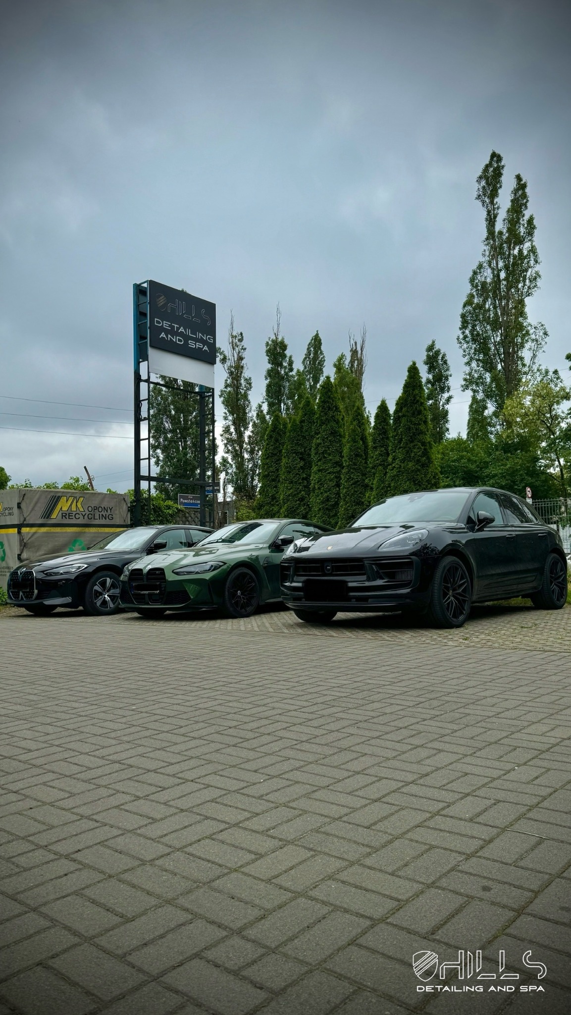 Rząd trzech samochodów: czarny BMW, zielony BMW M4 i czarny Porsche Macan, stojących na brukowanym parkingu przed salonem detailingowym Hills Detailing and Spa pod pochmurnym niebem.