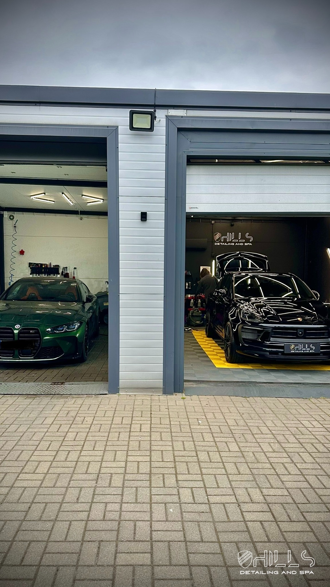 Widok z zewnątrz na dwa stanowiska detailingowe: zielony BMW M4 i czarny Porsche Macan w trakcie zabiegów pielęgnacyjnych. W tle widoczne logo firmy.