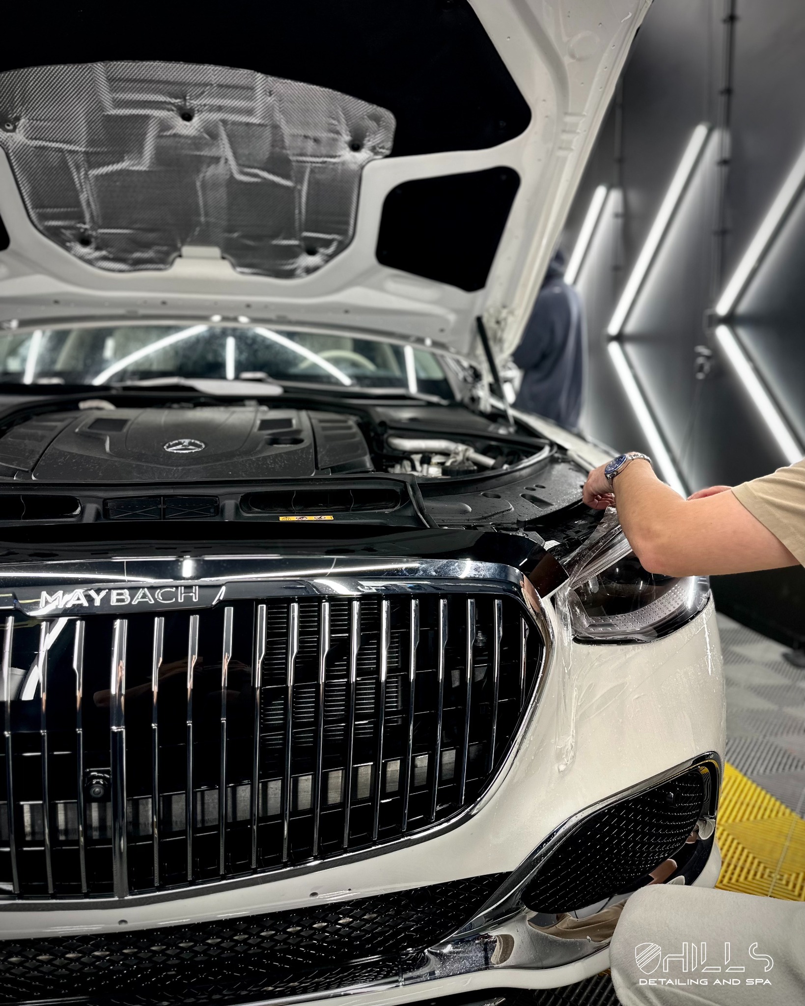 Biały Maybach z otwartą maską, w trakcie zabezpieczania folią ochronną przedniej lampy przez pracownika w warsztacie detailingowym z oświetleniem LED.