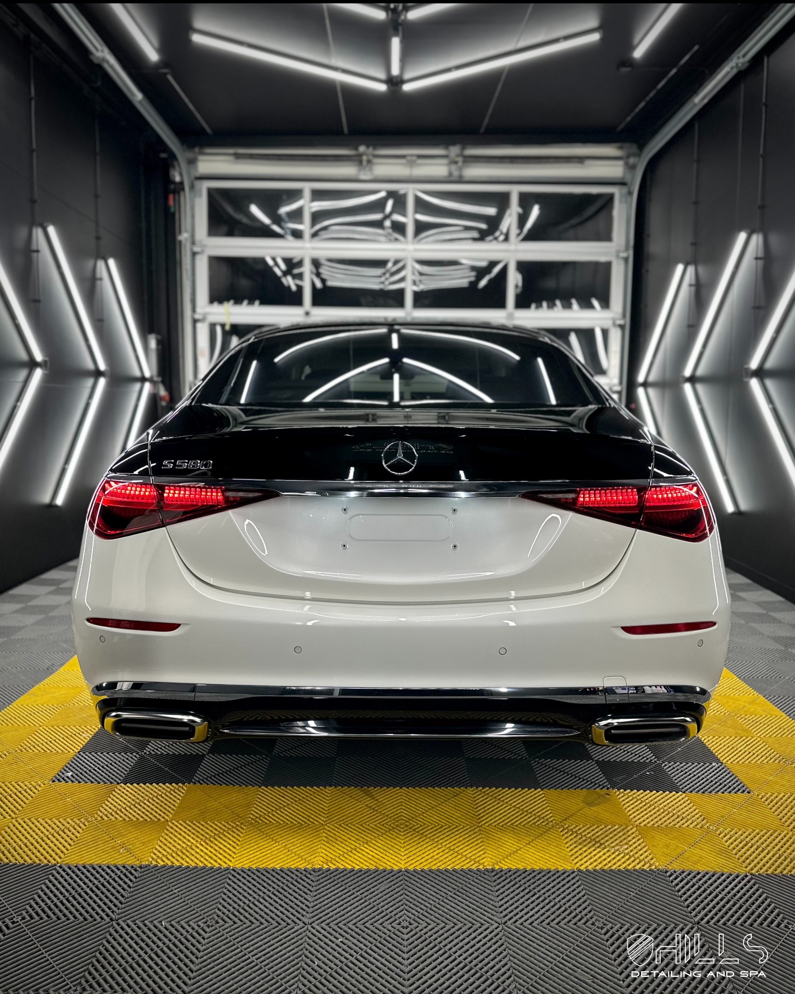 Tył dwukolorowego (czarno-białego) samochodu Maybach S580 zaparkowanego w dobrze oświetlonym garażu z podłogą w żółto-szarą szachownicę, widoczne tylne światła i logo marki.
