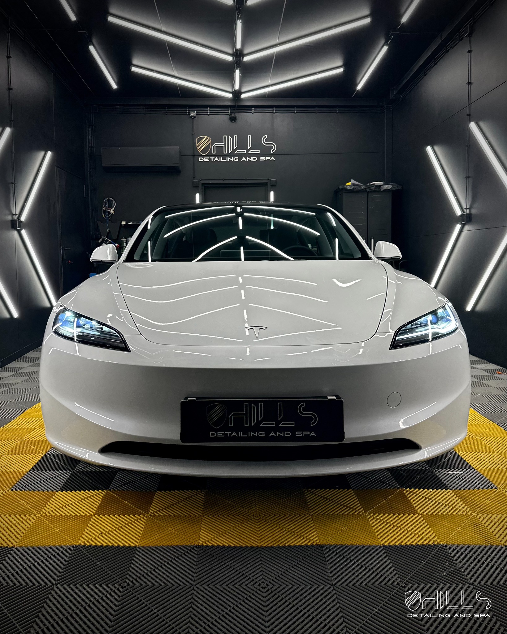 Biała Tesla Model 3 po zabiegach pielęgnacyjnych w sterylnym, oświetlonym studiu detailingowym z logo firmy na tablicy rejestracyjnej i podłodze w żółto-czarną kratę.