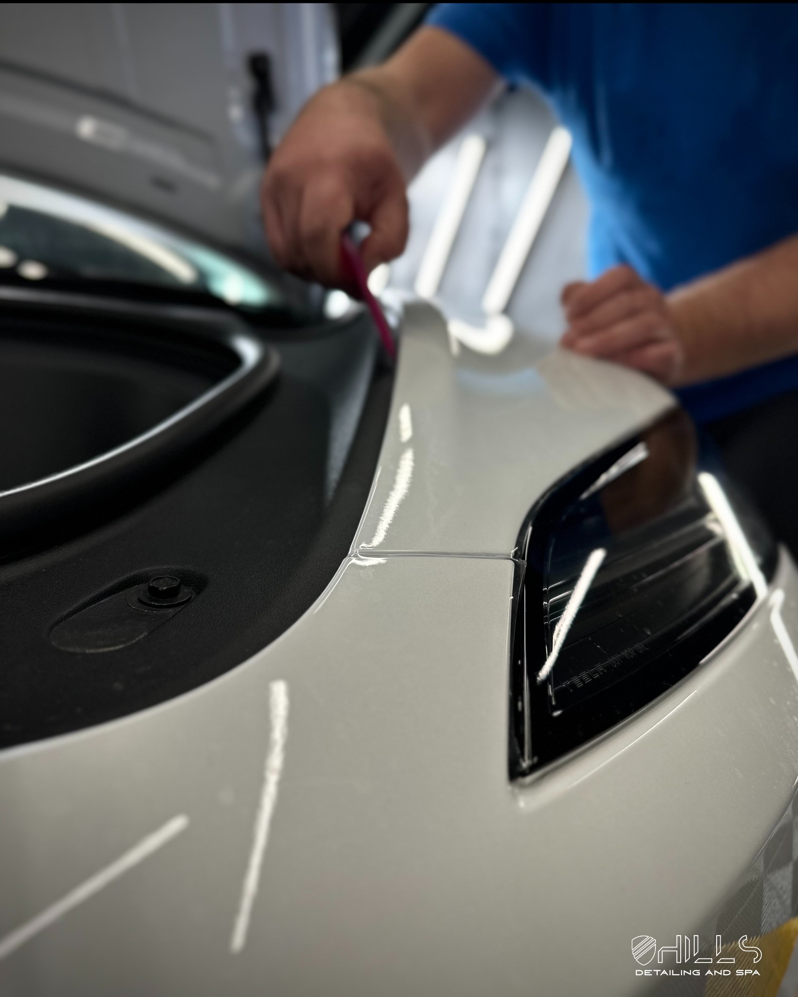 Aplikacja folii ochronnej na przednią część białego samochodu, widoczny fragment reflektora i narzędzie do aplikacji w ręce pracownika.