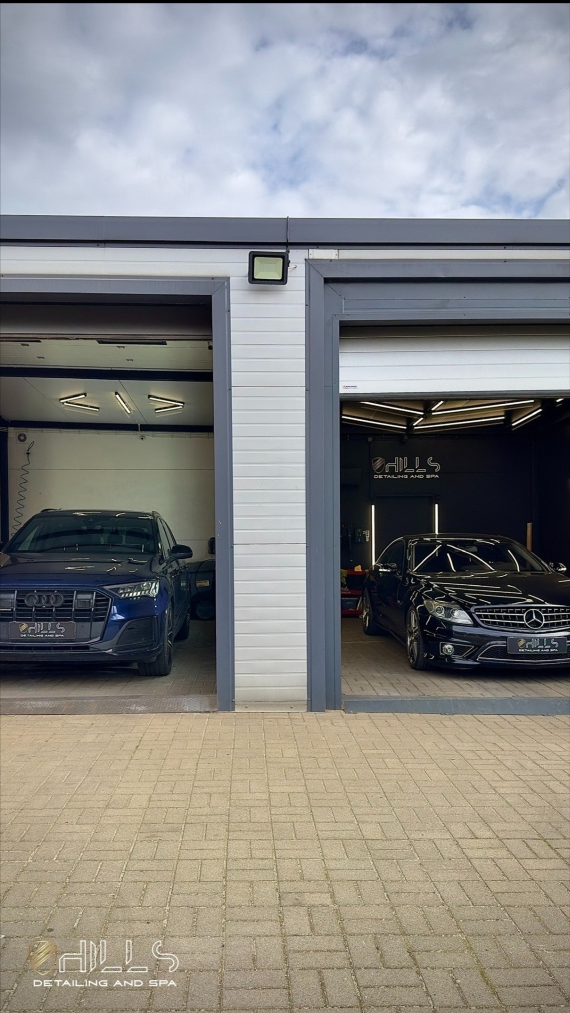 Dwa samochody, Audi i Mercedes, zaparkowane w otwartych garażach myjni detailingowej z logo firmy na ścianie i tablicach rejestracyjnych.