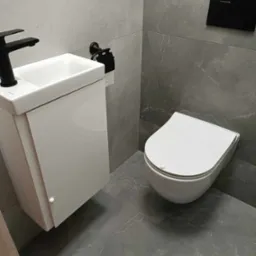 Nowoczesna łazienka z szarymi płytkami imitującymi marmur, wisząca toaleta i umywalka z szafką w kolorze białym, czarna bateria i akcesoria toaletowe.